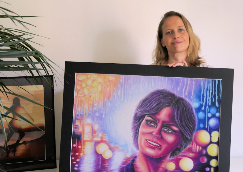 A photo showing the female artist standing next to her completed "Purple Rain" artwork, which is a highly detailed and vibrant colored pencil drawing.