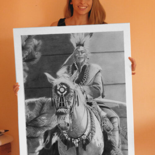 A photograph of an artist holding a large, photorealistic pencil drawing. The drawing is a highly detailed, black-and-white portrait of the elderly Native American actor Silkirtis Nichols (Buffalo Child) riding a horse, capturing his expressive face and traditional attire with precise shading.
